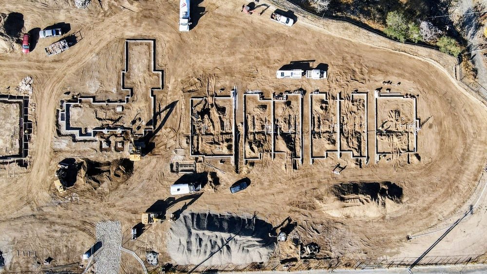 Aerial view of an apartment construction site with excavated foundation outlines, equipment, and work vehicles.