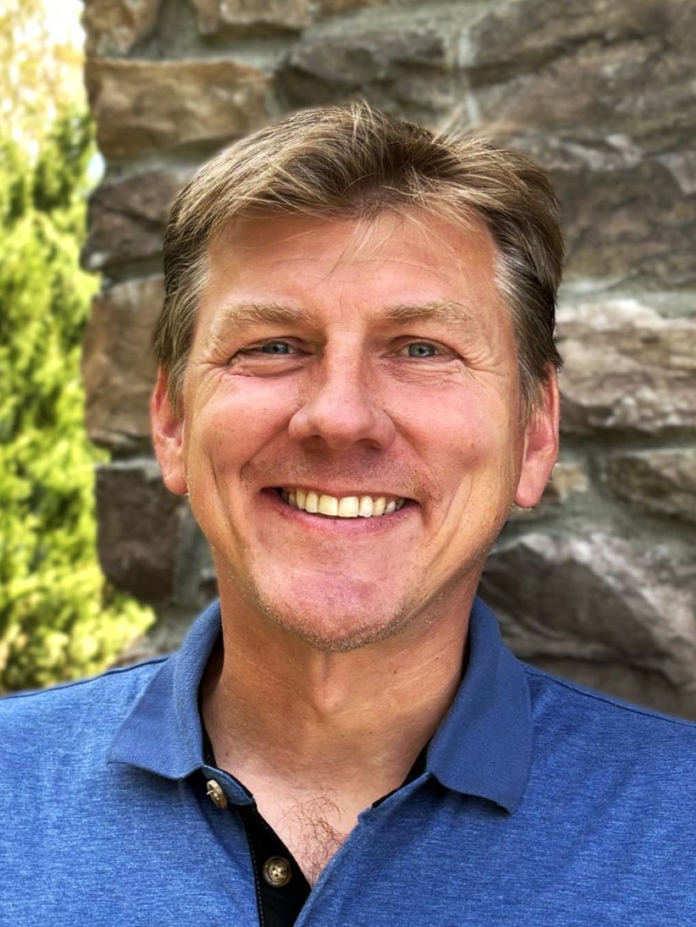 Headshot of Chris smiling outdoors in a blue polo shirt with a stone wall in the background.