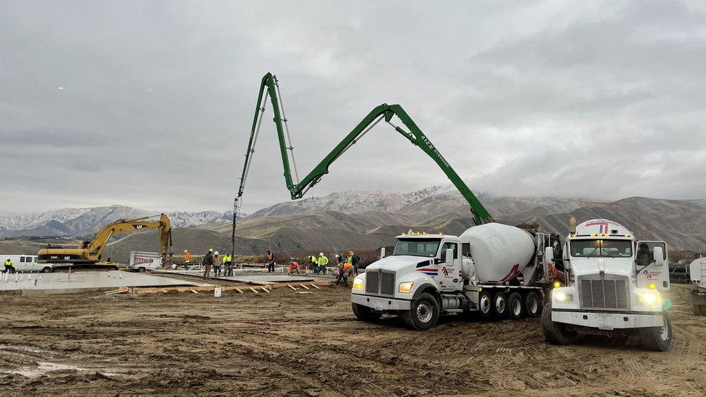 Concrete trucks and a pump boom supporting a foundation pour at a construction site with mountains in the background.