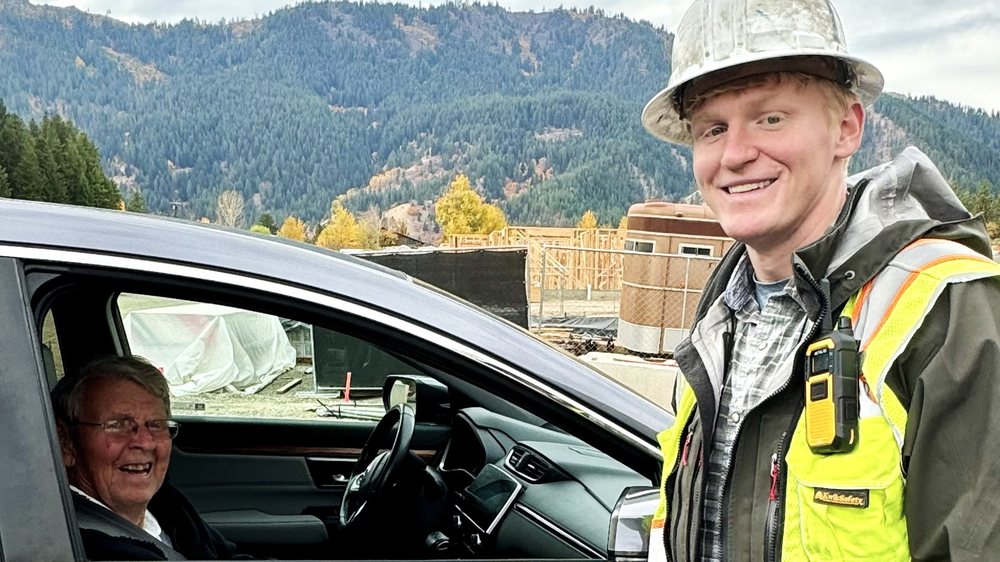 Construction manager wearing a hard hat and safety vest greeting a resident beside a car with forested mountains in the background.