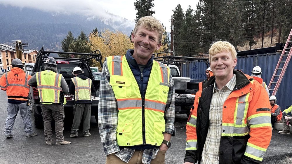 Two construction managers wearing safety vests standing together at an active jobsite with crew members in the background.