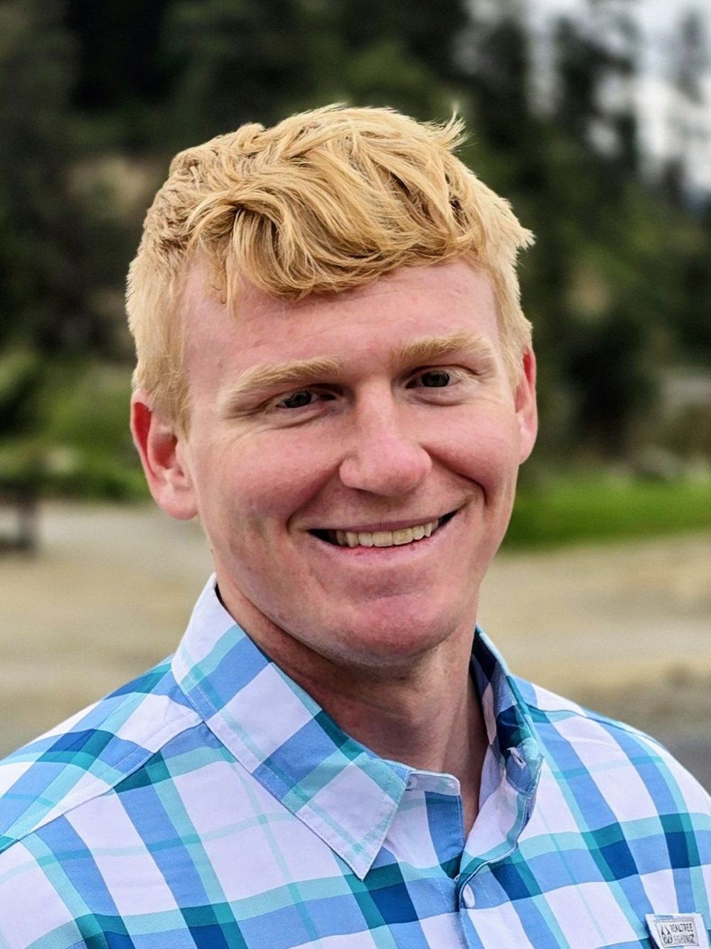 Headshot of Elliot smiling outdoors in a blue and white plaid shirt.