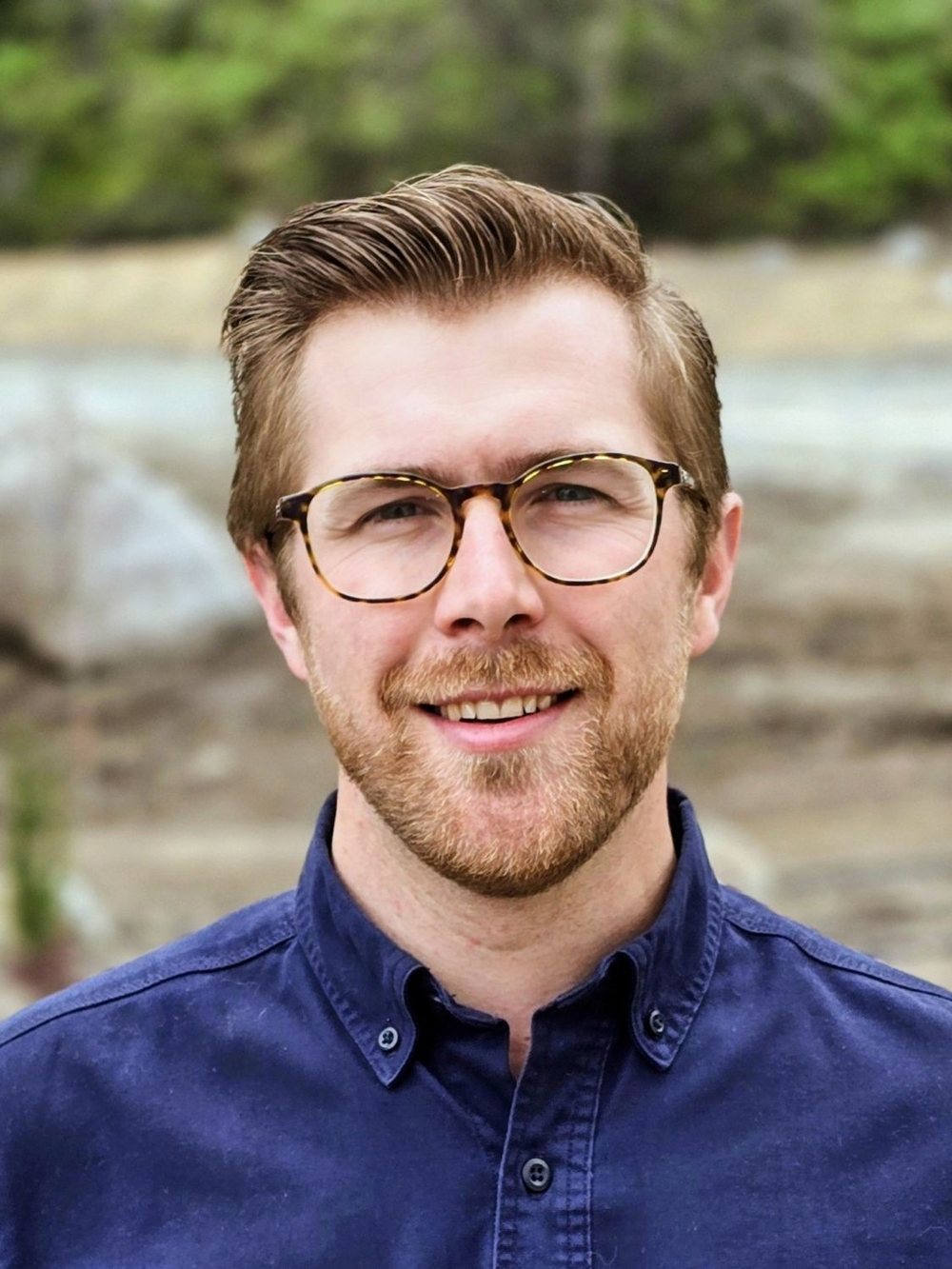 Headshot of Joshua R smiling outdoors in glasses and a navy button-down shirt.