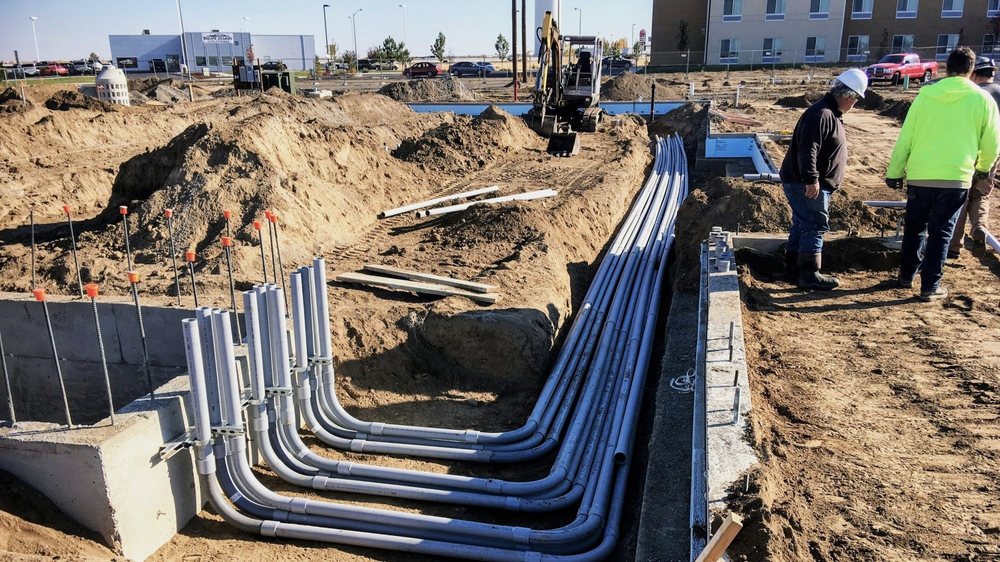 Underground utility conduits routed through a construction site trench beside foundation walls and excavation equipment.