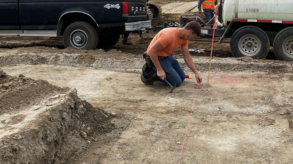Construction worker kneeling on a jobsite while marking a foundation layout line beside work vehicles and equipment.