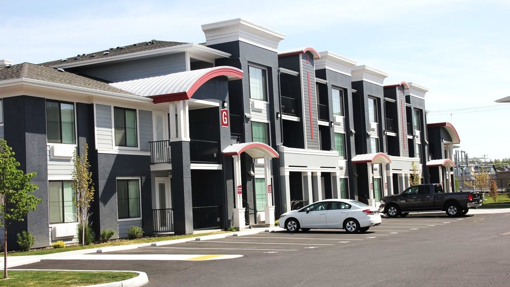 Modern apartment building with dark gray siding, red accents, covered entryways, and a parking lot.