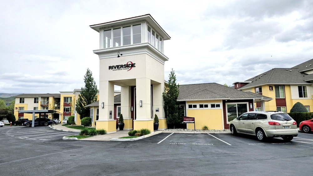 Riverside Apartment Homes leasing office with a tower entrance, landscaping, and nearby apartment buildings.