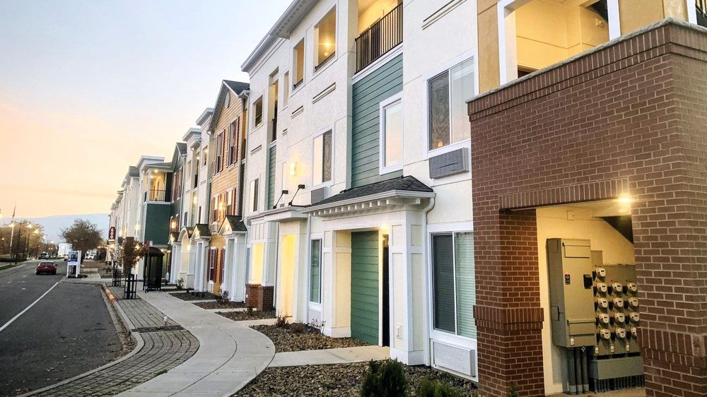 Riverside apartment homes and sidewalk along a city street at dusk with exterior lights glowing.